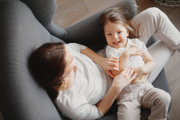 mother with a 3-year-old daughter sit in a gray armchair, hug and laugh - Valokuva, kuva