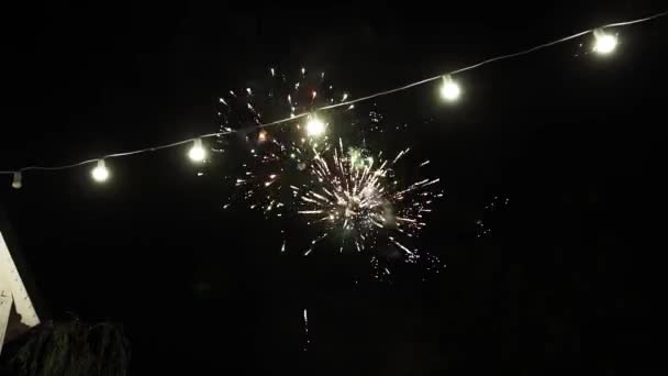 電球からの明るい光を背景に、夜遅くに夜空に美しく大きなカラフルな花火。パワフルなシェークと輝く色 - 映像、動画