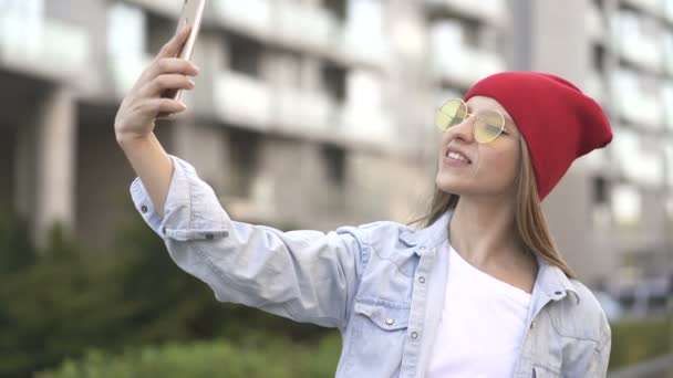 4k. Jeune jolie fille en casquette rouge et lunettes utilisent smartphone comme selfie dans la rue de la ville
.  - Séquence, vidéo