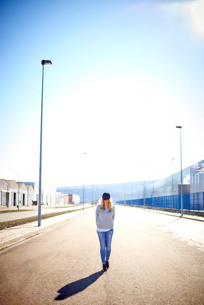 Menina com boné e cabelo loiro longo, vestindo jeans, tênis e camisola, andando pela rua em uma área urbana vazia em um dia muito ensolarado
 - Foto, Imagem