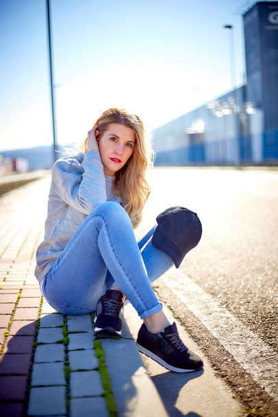 Girl with cap and long blonde hair, wearing jeans, sneakers and a sweatshirt, sitting on the sidewalk in an empty urban area on a very sunny day - Photo, Image