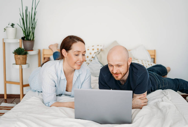 Adult married couple in casual clothing using open laptop lie on the bed at bright interior - Fotó, kép