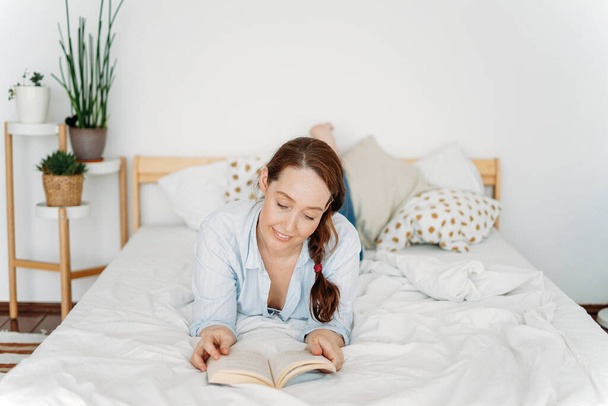 Ystävällinen viehättävä brunette nainen lukeminen kirja sängyllä valoisa sisustus
 - Valokuva, kuva