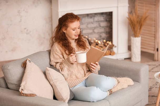 Όμορφη νεαρή γυναίκα πίνοντας ζεστό κακάο και διαβάζοντας το βιβλίο στο σπίτι - Φωτογραφία, εικόνα