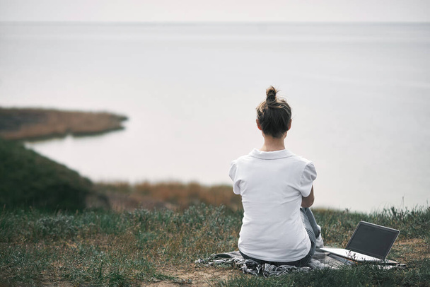mujer joven con el pelo rubio en camisa blanca se sienta en la colina cerca de la orilla del mar con el ordenador portátil y mirando delante de ella
 - Foto, Imagen