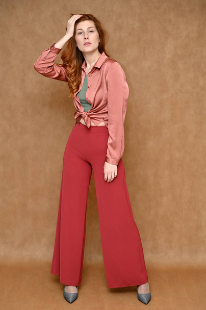 Nice girl model dressed in red pants and a pink blouse. Full-length vertical portrait of a young Caucasian red-haired woman on a beige background in various poses. - Fotó, kép