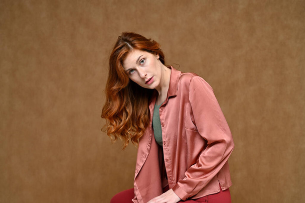 A pretty model with long hair is dressed in red trousers and a pink blouse. Portrait of a young Caucasian red-haired woman sitting on a stand on a beige background in various poses. - Φωτογραφία, εικόνα