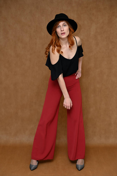 Full-length portrait of a pretty red-haired young woman in a black blouse, red trousers. Girl Model posing in the studio on a beige background with a hat in her hands. - Zdjęcie, obraz
