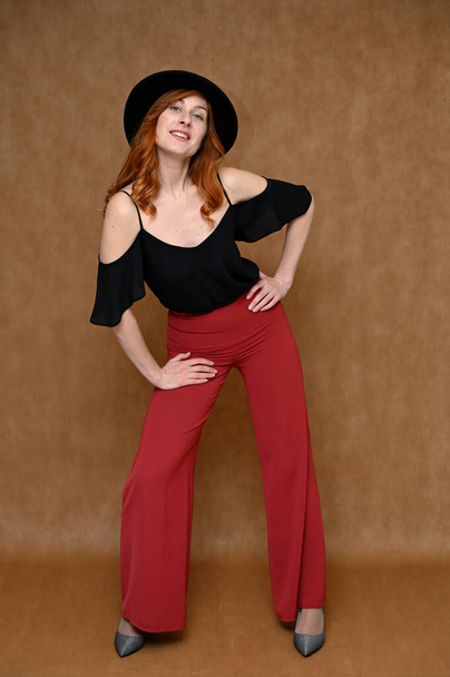 Full-length portrait of a pretty red-haired young woman in a black blouse, red trousers. Girl Model posing in the studio on a beige background with a hat in her hands. - Photo, image