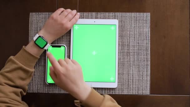 茶色のセーターの女の子は、キッチンのテーブルに座っている間、緑色の画面を持つスマートガジェットを使用します - 映像、動画