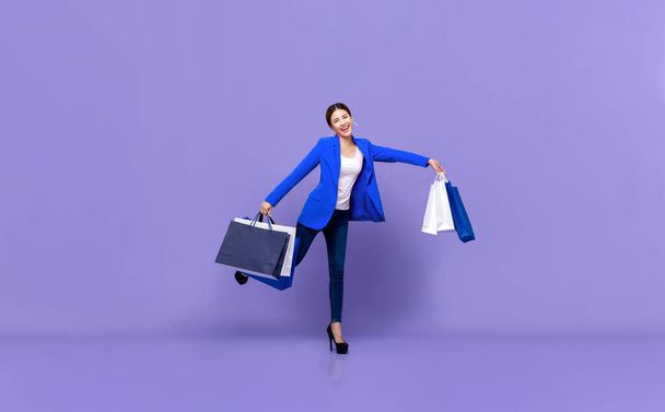 Sonriente hermosa mujer asiática joven en ropa blanca azul sosteniendo bolsas de compras de pie en el fondo púrpura
 - Foto, Imagen