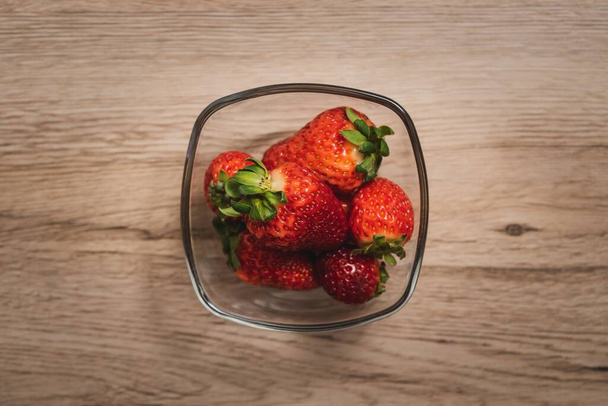 An overhead shot of strawberries in a glass bowl on a wooden background - Foto, Imagen