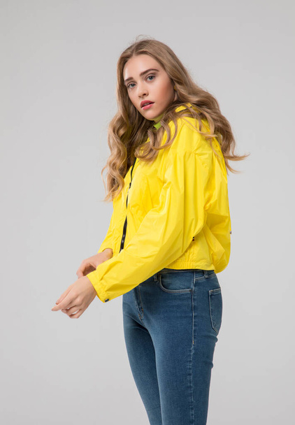 blonde girl posing in a yellow raincoat and blue denim jeans. in studio on white background. - 写真・画像