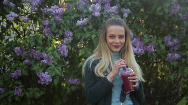 Attractive girl with long hair dressed in a dark emerald green coat is drinking a smoothie - Imágenes, Vídeo