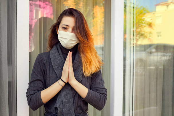 Άρρωστη γυναίκα που μένει στο σπίτι και προσεύχεται για προστασία από τον κορωνοϊό φορώντας μια ιατρική μάσκα μιας χρήσης στο πρόσωπό της. - Φωτογραφία, εικόνα