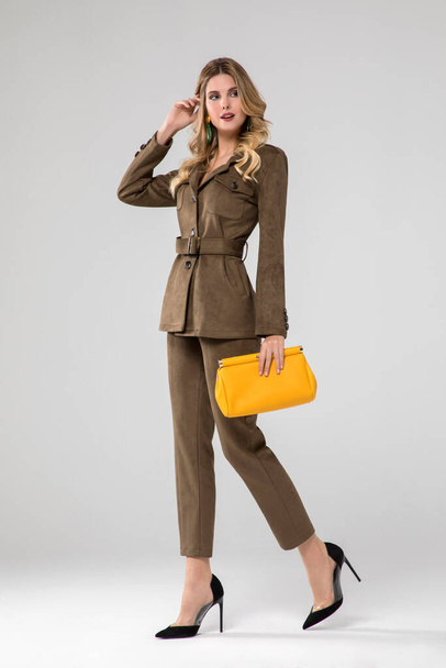 hinreißend blondes Model posiert in einem olivgrünen Lederanzug. Gelbe Handtasche. Stehen. weißer Hintergrund. Studioaufnahme. - Foto, Bild