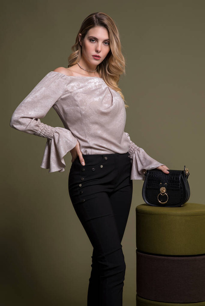 hinreißend stylisches blondes Model posiert in weißer Bluse und schwarzer Hose, High Heels und schwarzer Tasche. Hand auf ihre Taille. Olivgrüner Hintergrund. - Foto, Bild