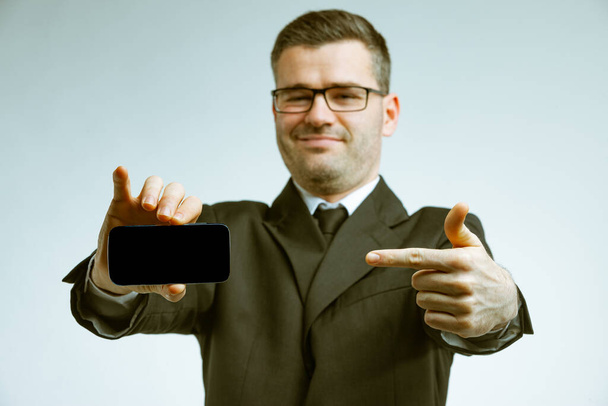 Νεαρός με κοστούμι και γυαλιά με ένα χαμόγελο στο πρόσωπό του, δείχνοντας με το δείκτη του τη μαύρη οθόνη ενός smartphone στο χέρι του. Χαμηλή γωνία εμπρός πορτρέτο σε καθαρό φόντο - Φωτογραφία, εικόνα