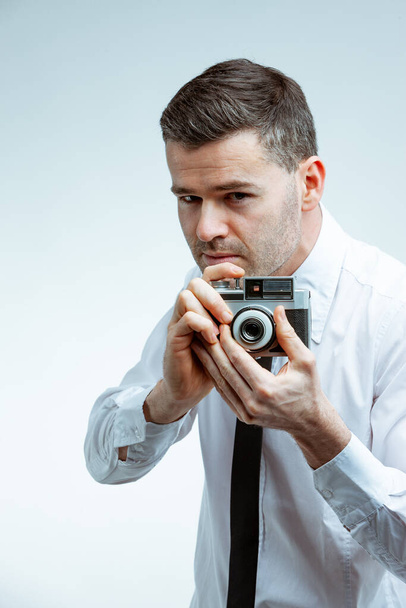 Όμορφος άνδρας σε λευκό πουκάμισο με μαύρη γραβάτα, τραβώντας μια φωτογραφία με vintage φωτογραφική μηχανή. Κοντινό πλάνο μπροστά πορτρέτο σε φωτεινό καθαρό φόντο - Φωτογραφία, εικόνα