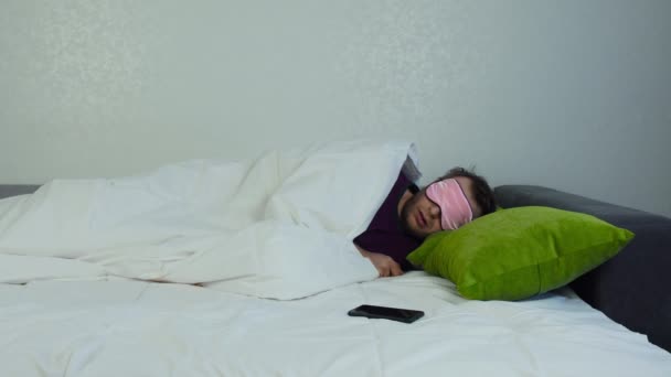 Man with blindfold and covered with blanket is asleep and he was awakened by sound of alarm clock on smartphone, guy turns off alarm, turns over on his side and continues to sleep. Laziness concept - Materiaali, video