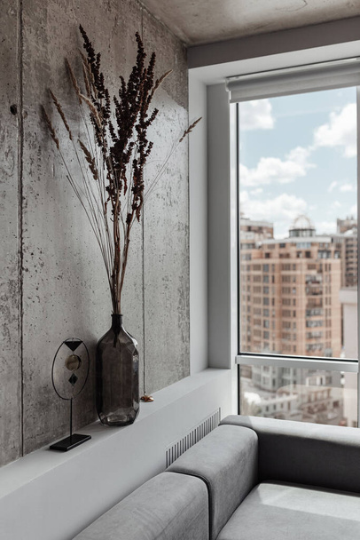 un fragmento del interior, un marco vertical con un sofá gris, un jarrón con flores secas y una caña cerca de una gran ventana panorámica con vistas a la ciudad
 - Foto, Imagen