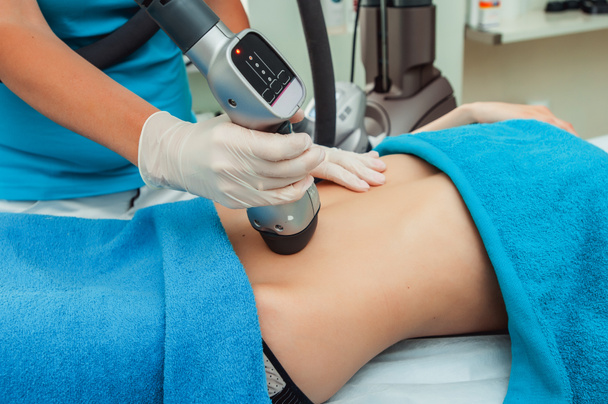 Cropped shot of a professional dermatologist in white gloves performing radiofrequency lifting procedure on the stomach of a woman in a blue towel - Fotografie, Obrázek