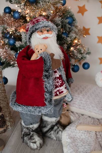 Mikulás baba, nagy karácsonyi stand figura gazdaság aranyos plüssmackó, elhelyezett előtt egy kivilágított karácsonyfa, ünnepi dekoráció téli ünnepekre, boldog idők ünneplő családi koncepció - Fotó, kép