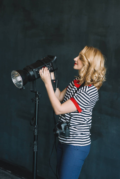 Νεαρή όμορφη φωτογράφος κορίτσι με μια φωτογραφική μηχανή στα χέρια της εργάζεται σε ένα στούντιο φωτογραφιών χρησιμοποιώντας τεχνητό φωτισμό. Η δουλειά των γυναικών είναι χόμπι. Μαλακή επιλεκτική εστίαση. - Φωτογραφία, εικόνα