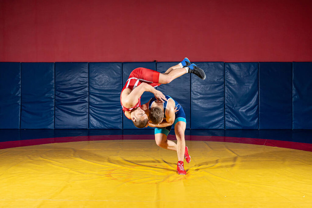 Két erős birkózó kék és piros birkózó harisnyában birkózik és csípődobást végez egy birkózó szőnyegen az edzőteremben. Fiatalember csimpaszkodik.. - Fotó, kép