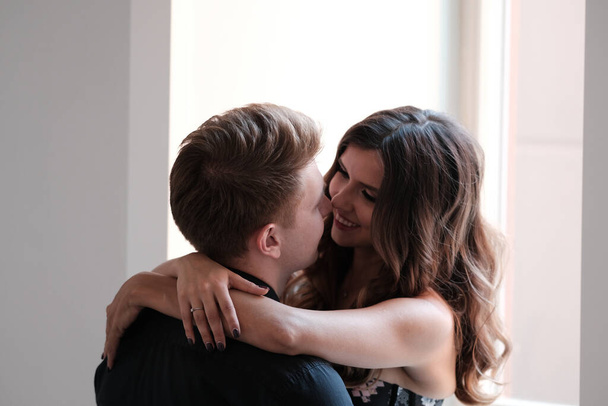 A young couple - a girl in a magnificent dress and a guy in a black shirt and trousers hug and kiss in a hotel room.  - Photo, image