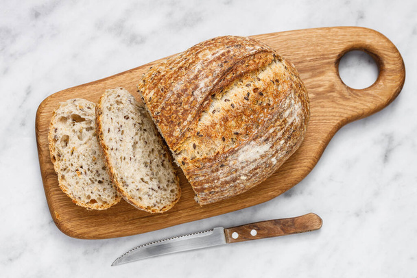 Round loaf of freshly baked sourdough bread with knife on cutting board, top view. Artisan bread with seeds on marble table. Rustic sourdough bread. - Photo, Image