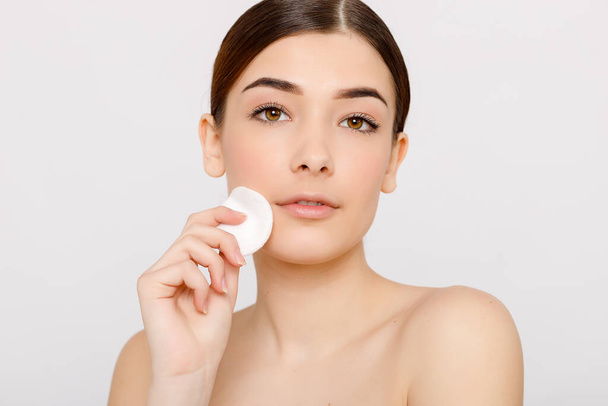 Healthy fresh girl removing makeup from her face with cotton pad. Beauty woman cleaning her face with cotton swab pad isolated on grey background. Skin care and beauty concept. - Zdjęcie, obraz