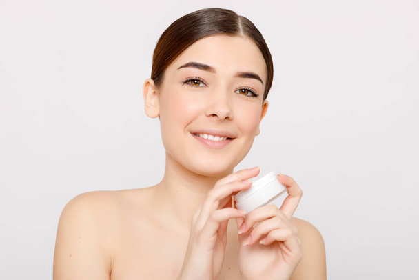 Beauty portrait of girl with perfect nude make-up. Holding cream, some cream on finger. Looking at camera and smiling. Beauty salon. Head and shoulders, studio, indoors - Photo, Image