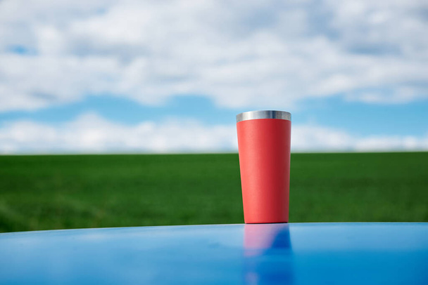 Close-up picture of red thermos cup with tea, standing on blue car top in front of green field and light blue sky with white clouds Picnic in countryside. Nature protection concept. - Photo, Image