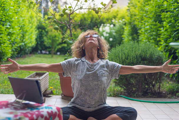 Зріла жінка з окулярами, що виконує позицію йоги з планшетом, медитація на відкритому повітрі в зеленому саду. Карантин Ковіда-19 залишається вдома. Рішення для блокування
. - Фото, зображення