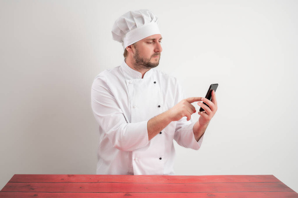 Portrait of young male chef in white uniform with blonde hair, holding smartphone, texting messages posing on a white isolated background - Photo, image