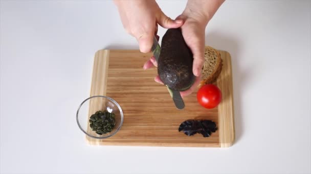 Girl cuts a ripe avocado. On a wooden board, a girl cuts a ripe avocado with a knife. On a white table are avocados, bread, pumpkin seeds, tomatoes, lettuce. Close-up. - Video