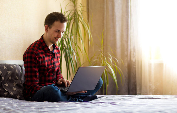 Młody człowiek wolny strzelec pracujący z laptopem w domu w łóżku przy oknie. Telepraca - Zdjęcie, obraz