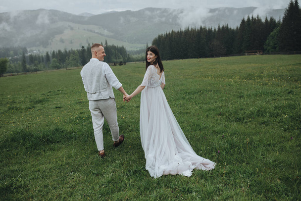 Spring wedding in the mountains. A guy in a shirt and vest and a girl in a white dress walk along a wooden fence along a green meadow against the backdrop of mountains and forests - Foto, Bild