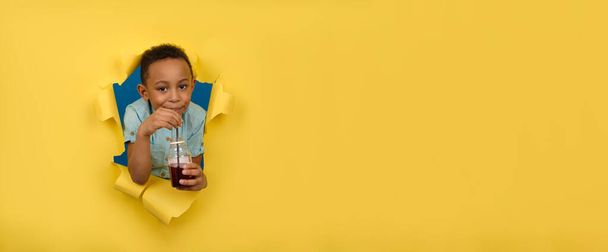 Happy African-American boy in blue shirt is holding juice bottle, drinking red cherry drink from black bar straw, against yellow background of torn paper wall. Healthy drink detoxification concept - 写真・画像