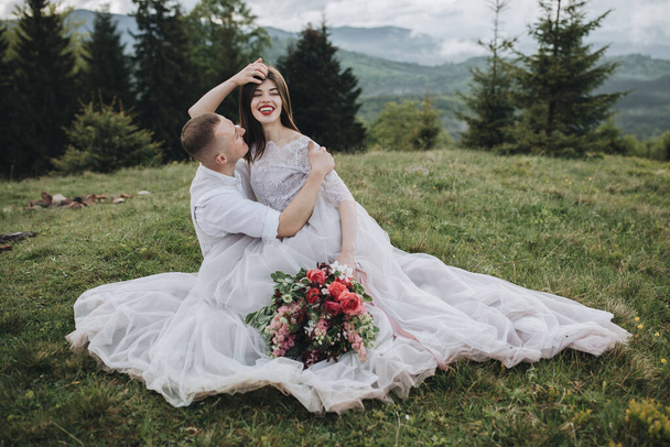 Spring wedding in the mountains. A young guy in a white shirt and trousers and a girl in a white dress are sitting on the green grass in the mountains and holding a bouquet of red flowers - Foto, Bild