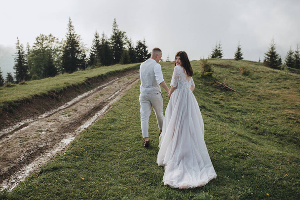 Spring wedding in the mountains. A guy in a shirt and vest and a girl in a white dress walk along a mountain road at sunset among the clouds - Fotó, kép