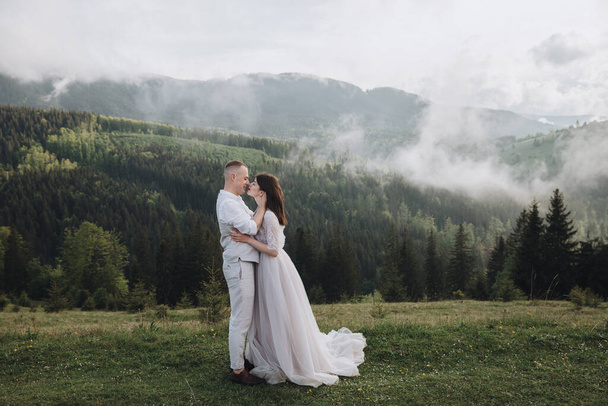 Spring wedding in the mountains. A young guy in a white shirt and trousers and a girl in a white dress stand on a mountain peak against the background of sky and trees - 写真・画像