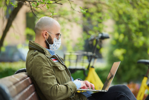 A man in a medical face mask to avoid the spread coronavirus works remotely on a laptop in the park. A guy wears a pilot sunglasses sits on a bench on the street with a computer near a bicycle. - Zdjęcie, obraz