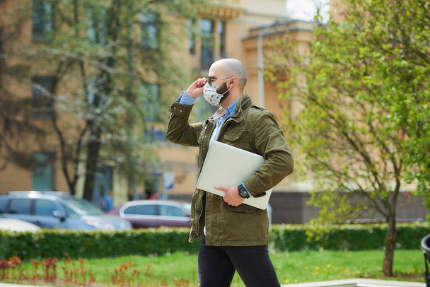 Un homme chauve avec une barbe dans un masque médical pour éviter la propagation du coronavirus marche avec un ordinateur portable dans le parc. Un mec porte un masque facial n95 et des lunettes de soleil pilotes dans la rue de la ville
. - Photo, image