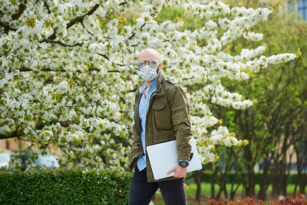 Un hombre calvo con barba en una mascarilla médica para evitar la propagación del coronavirus camina con un portátil en el parque. Un tipo usa mascarilla n95 y gafas de sol piloto en el jardín entre árboles florecientes
. - Foto, imagen