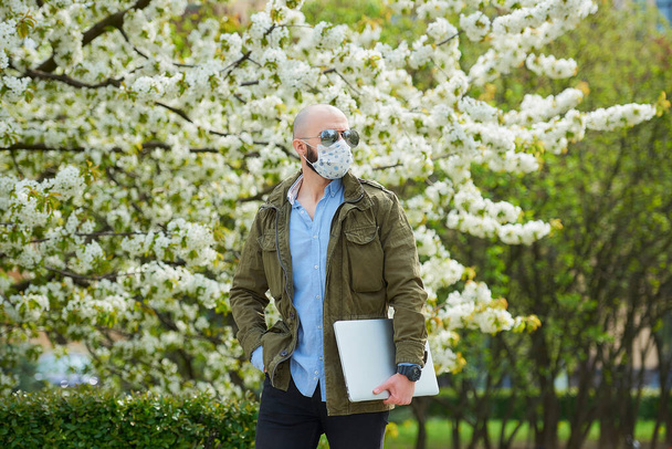 Ένας φαλακρός άντρας με γενειάδα σε μάσκα ιατρικού προσώπου για να αποφύγει την εξάπλωση του coronavirus περπατάει με ένα φορητό υπολογιστή στο πάρκο. Ένας τύπος φοράει μάσκα προσώπου N95 και γυαλιά ηλίου στον κήπο ανάμεσα σε ανθισμένα δέντρα.. - Φωτογραφία, εικόνα