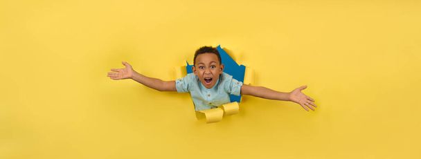Cheerful and happy African-American on torn yellow paper wall background reaching out to support or take something asks to be picked up or hugged, boy asks to be picked up joyfully waving his arms - Zdjęcie, obraz