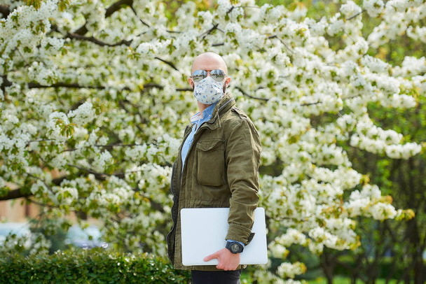 Ein glatzköpfiger Mann mit Bart und medizinischer Gesichtsmaske zur Vermeidung der Ausbreitung des Coronavirus spaziert mit einem Laptop durch den Park. Ein Mann trägt eine Gesichtsmaske und eine Pilotensonnenbrille im Garten zwischen blühenden Bäumen. - Foto, Bild