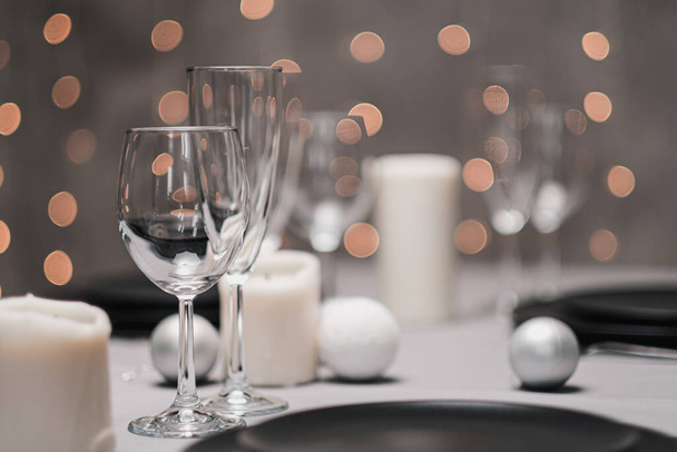 A table set in the restaurant. Serving a table. Grey artistic background. Black matte utensils, glass glasses. White candles and decor. Festive yellow lights garland in the background. - Фото, изображение